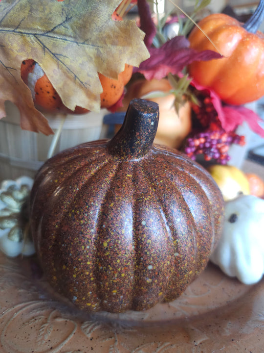 A decorative chocolate pumpkin in a mottled brown and orange color with a dark stem, displayed on a table with autumn leaves and other pumpkins in the background.