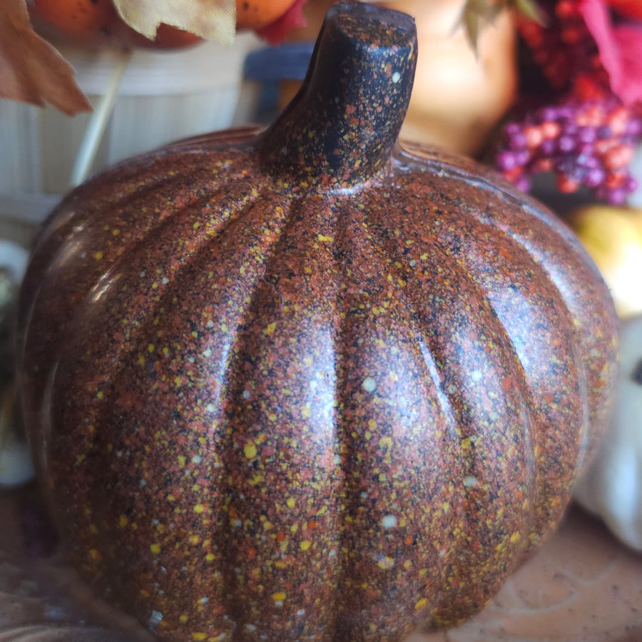 A decorative chocolate pumpkin in a mottled brown and orange color with a dark stem, displayed on a table with autumn leaves and other pumpkins in the background.